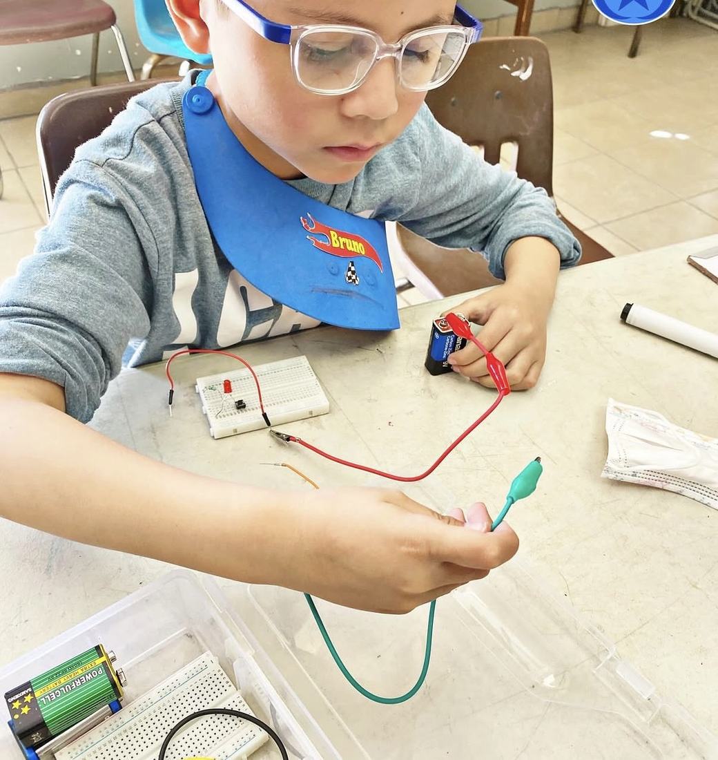 Niño de primaria armando su primer circuito con una pila, cables y un foco en un taller de Qubiko