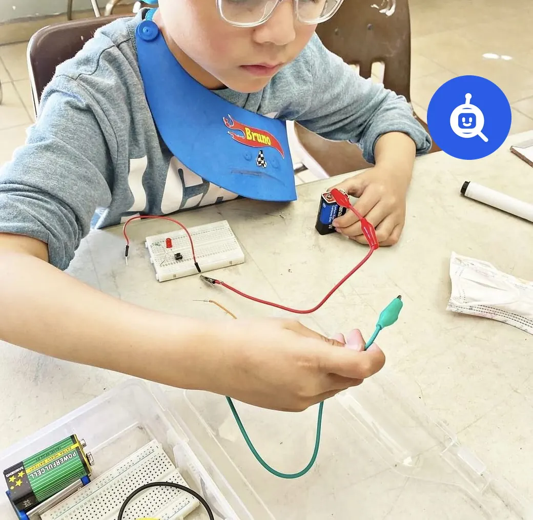 Niño de primaria armando su primer circuito con una pila, cables y un foco en un taller de Qubiko