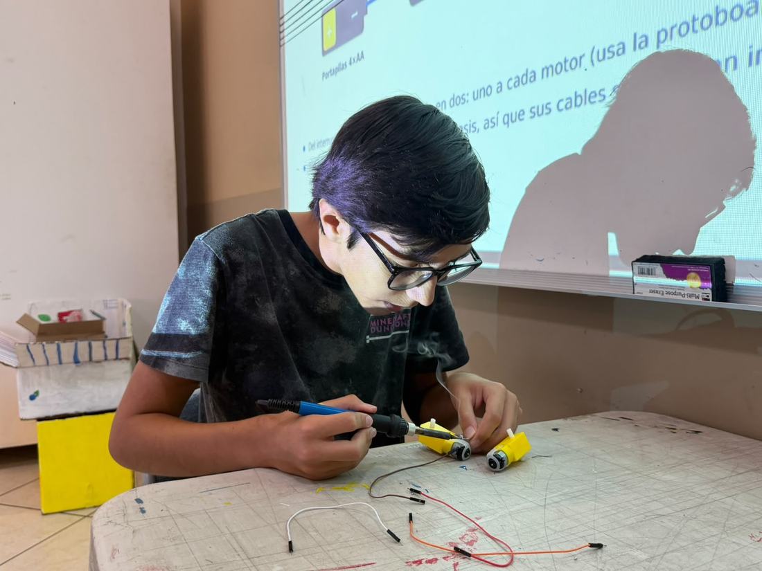 Alumno de secundaria arma un robot móvil con protoboard y motores durante un taller de Qubiko, con la clase proyectada al fondo
